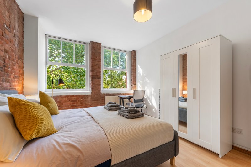 Bright bedroom with double bed, mustard cushions, exposed brick wall, large windows and white wardrobe