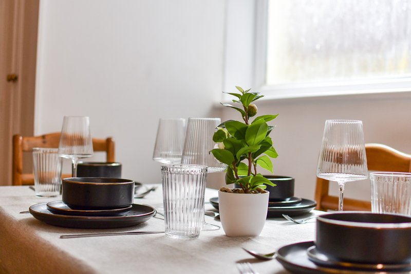 Dining table set with dark plates, glasses, and potted plant centerpiece by window