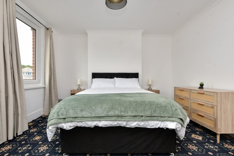 Bright bedroom with double bed, green bedcover, wooden dresser, and window with curtains