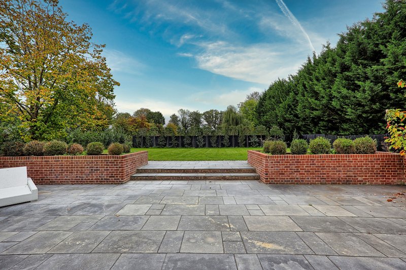 Brick garden wall with paved patio, manicured lawn and mature trees beyond