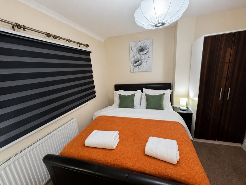 Bedroom with double bed, orange throw, striped window blinds, dark wardrobe and ceiling light