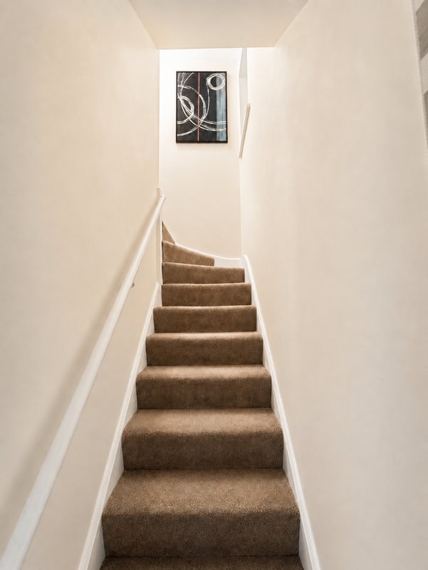 Narrow staircase with brown carpet, white walls and handrail, framed artwork above