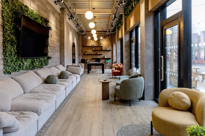Modern lounge with long grey sofa, curved armchairs, wooden table, and large windows overlooking city street