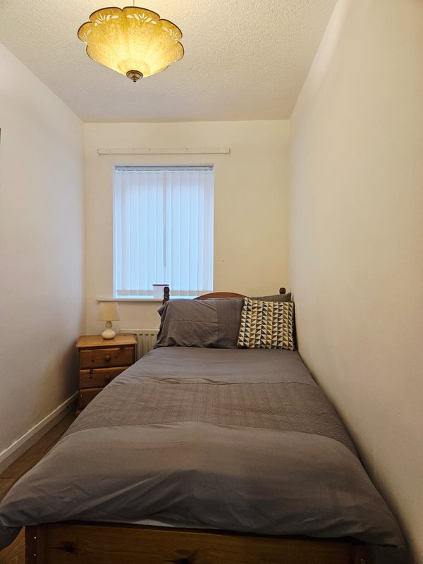 Small bedroom with single bed, wooden nightstand, ceiling light and vertical blinds