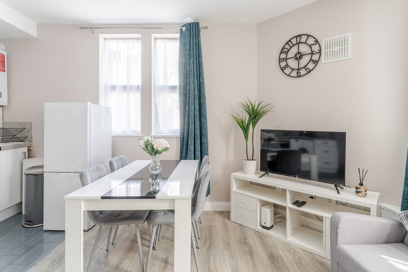 Open-plan living area with white dining table, chairs, TV unit, and kitchen appliances
