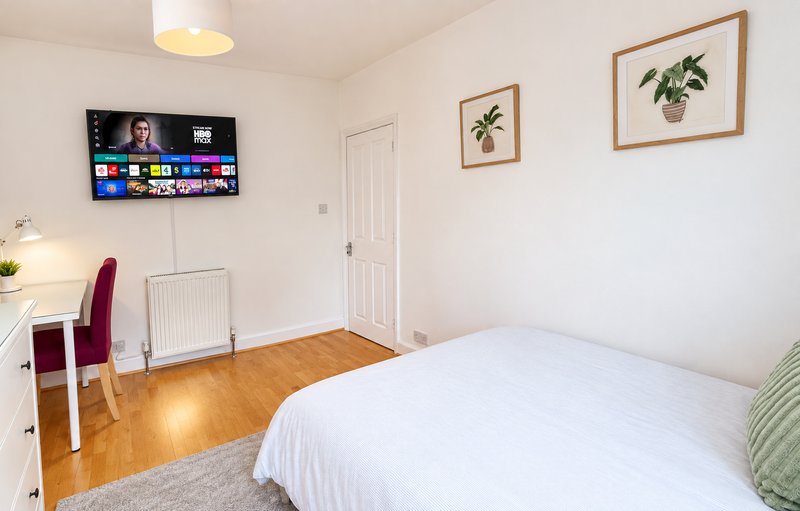 Bright bedroom with double bed, TV on wall, desk with chair, wooden floor and plant artwork
