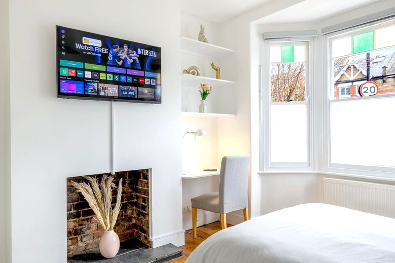 Bright bedroom with wall-mounted TV, fireplace with decorative vase, wooden desk chair and bed