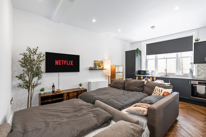 Open-plan living room with grey corner sofa, wall-mounted TV, kitchen area, and wooden flooring