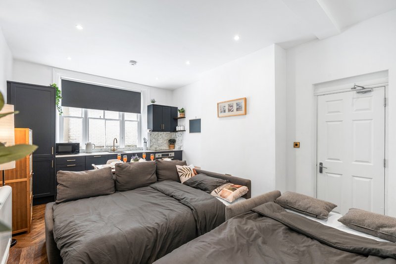 Modern bedroom with two beds, dark bedding, kitchenette, and window with black blinds