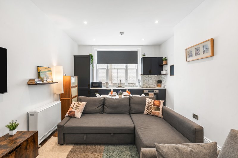 Open-plan living room with gray sectional sofa, black kitchen units, and wooden furniture