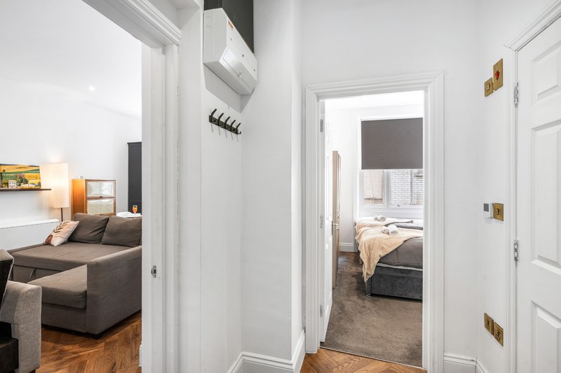Modern apartment hallway with white walls, wooden floor, and view into furnished bedroom and living room