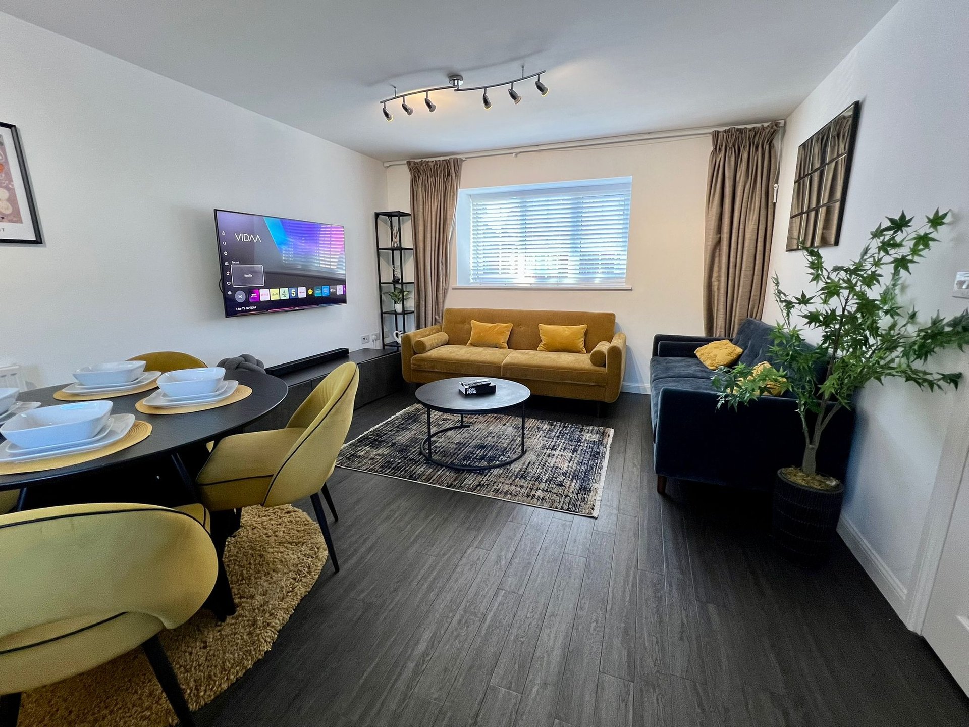Open-plan living and dining area with mustard sofa, dining table, TV, and potted plant