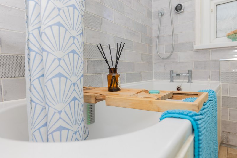 Modern bathroom with white bathtub, wooden shelf, blue patterned shower curtain and grey tile walls