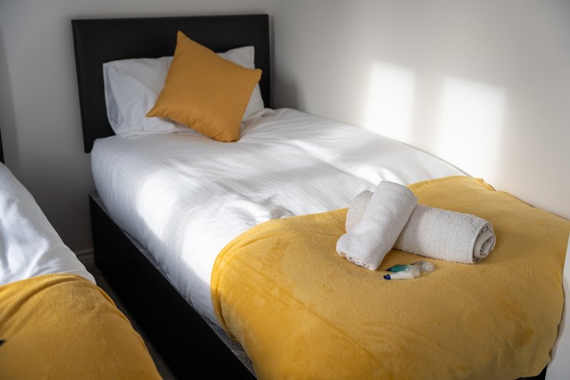 Single bed with black headboard, white sheets, yellow cushion and throw blanket