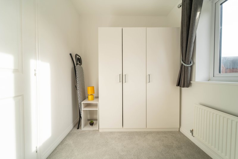 Bright bedroom with white built-in wardrobe, carpet flooring, radiator and window with curtains