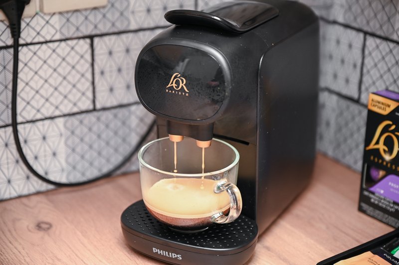 Black coffee maker with cup on wooden counter, kitchen appliances visible