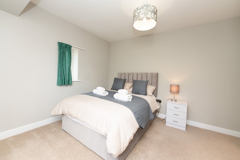 Bright bedroom with double bed, grey headboard, white bedside table and green curtains