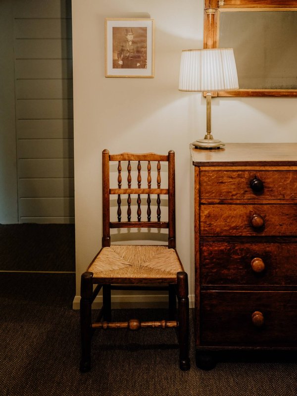 Bedroom corner with wooden chair, dresser, table lamp, framed artwork and mirror