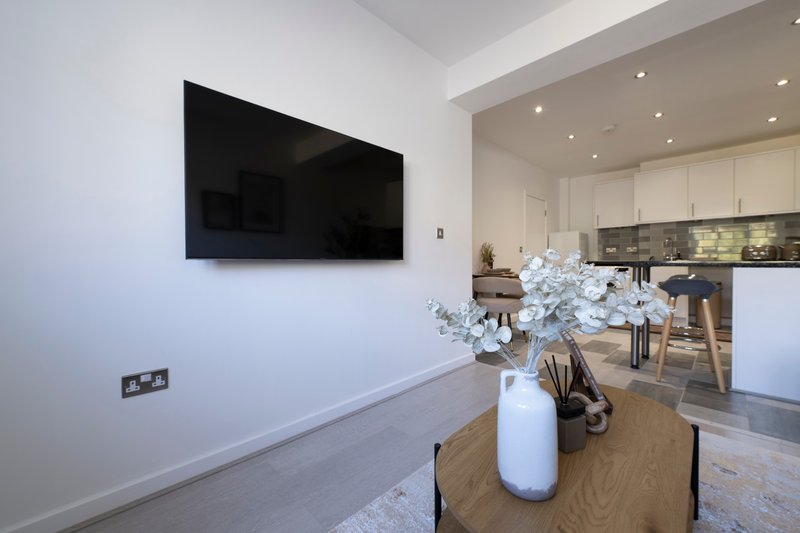 Modern open-plan living space with wall-mounted TV, wooden dining table, white flower arrangement, and kitchen visible in background
