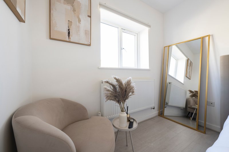 Bright modern bedroom with curved beige chair, decorative pampas grass, large mirror and window