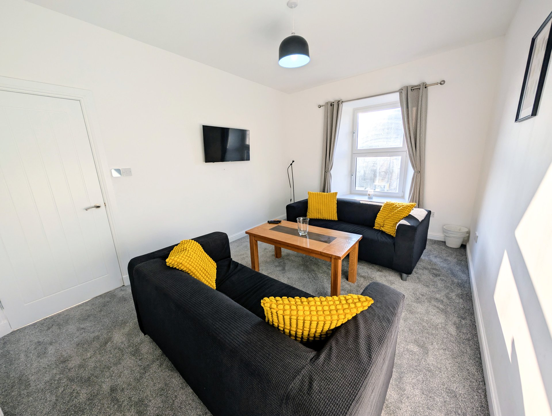 Bright living room with two black sofas, wooden coffee table, yellow cushions, wall-mounted TV, and window with curtains