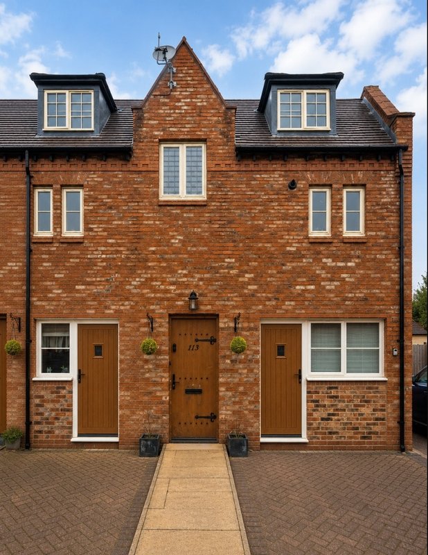 Red brick two-storey house with black roof, dormer windows, central brown door and paved driveway