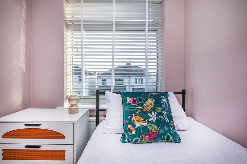 Pink bedroom with single bed, white pillows, patterned cushion, nightstand and window shutters