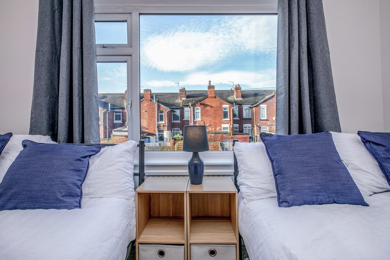 Bedroom with double bed, blue cushions, wooden bedside table, and large window overlooking brick buildings