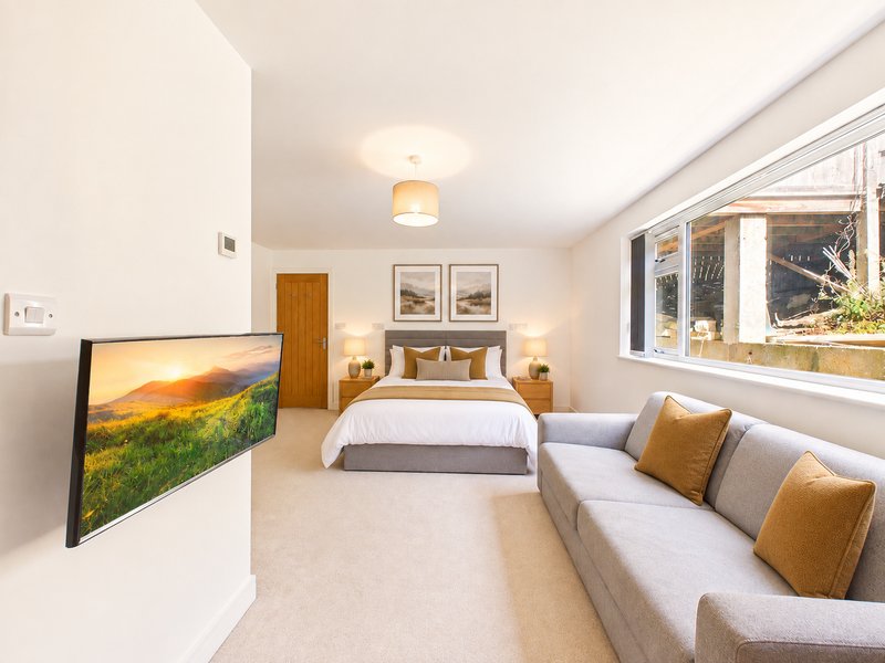 Spacious bedroom with double bed, grey sofa, wall-mounted TV and large windows overlooking garden