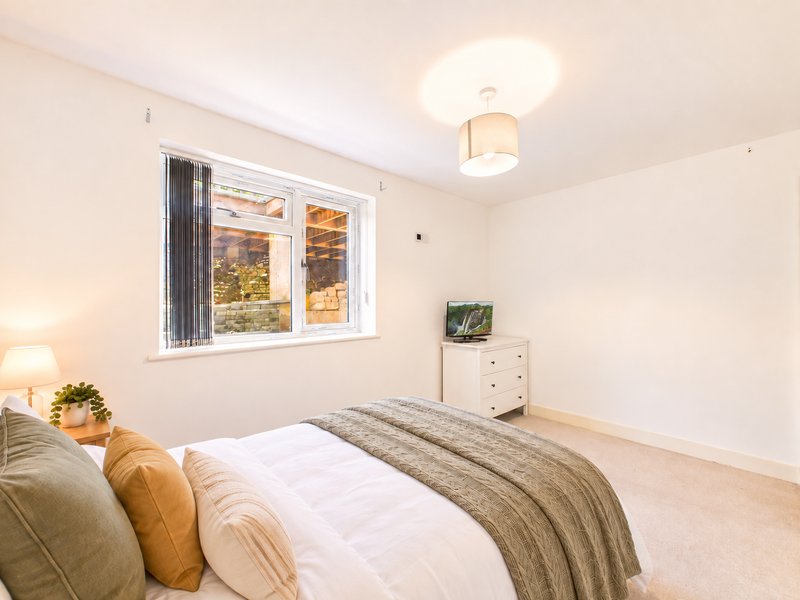 Bright bedroom with double bed, white bedding, striped throw, cushions, wooden bedside table with lamp, and window with garden view