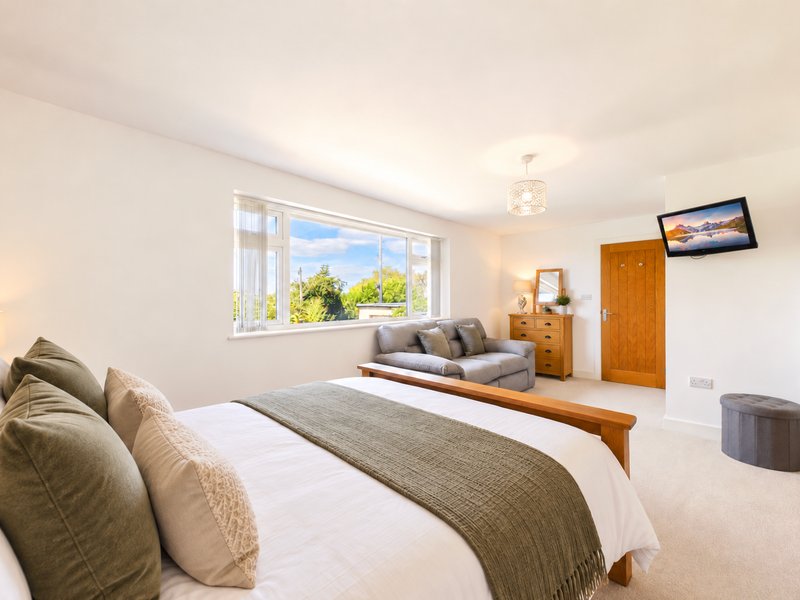 Bright bedroom with double bed, wooden headboard, cushions, seating area and large window with garden view