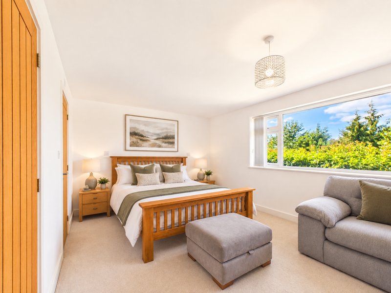 Bright bedroom with wooden bed, grey upholstered seating, and large windows overlooking garden