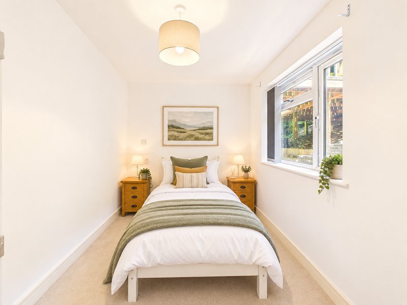 Bright bedroom with single bed, wooden bedside tables, framed landscape artwork, and windows