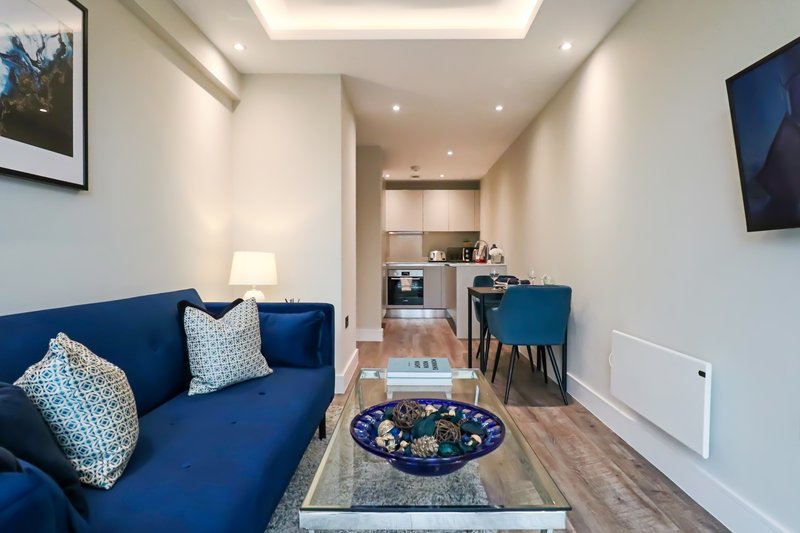 Modern open-plan living room with blue sofa, wooden floor, and kitchen visible in background