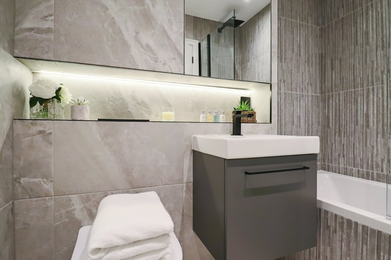 Modern bathroom with grey tiled walls, floating vanity, mirror with shelf lighting, and white towels