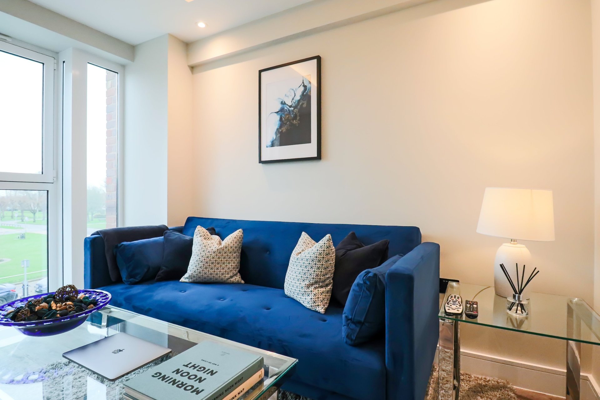 Bright living room with blue velvet sofa, decorative cushions, glass coffee table, and garden view