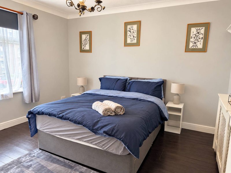 Bright bedroom with double bed, blue bedding, white nightstands, framed botanical prints, and wooden flooring
