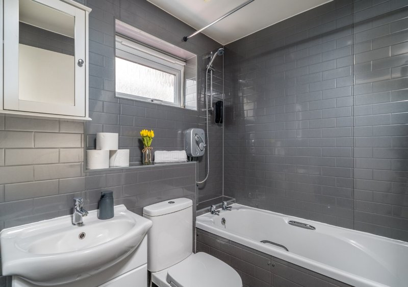 Modern bathroom with gray tile walls, white sink, toilet, bathtub with shower screen and yellow flowers