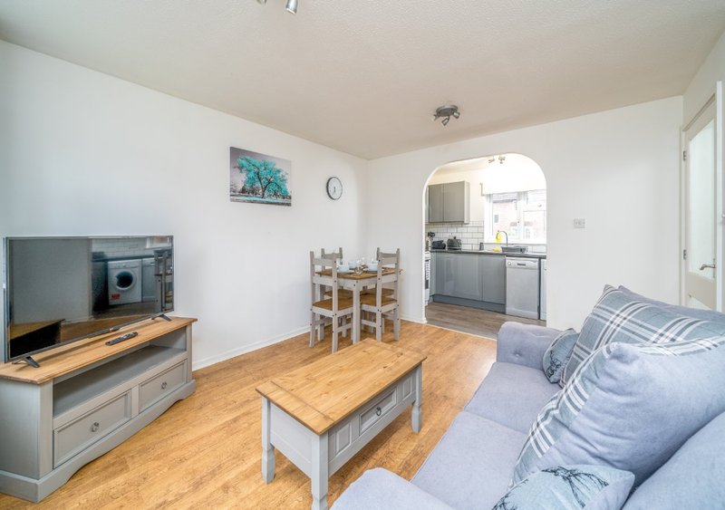 Open-plan living room with sofa, wooden furniture, TV, and kitchen area visible beyond