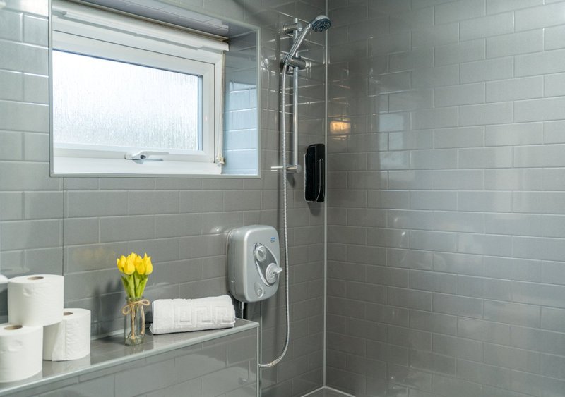 Modern bathroom with walk-in shower, grey tile walls, window, and shelf with flowers