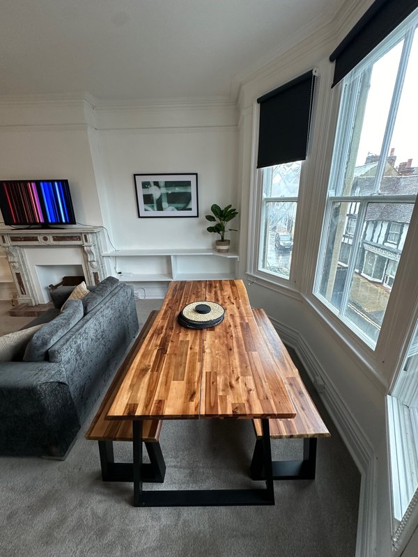 Living room with wooden dining table, gray sofa, fireplace, framed artwork, and large windows with city view