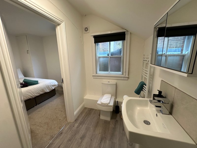 Bathroom with toilet, sink, mirror and window; bedroom visible through doorway