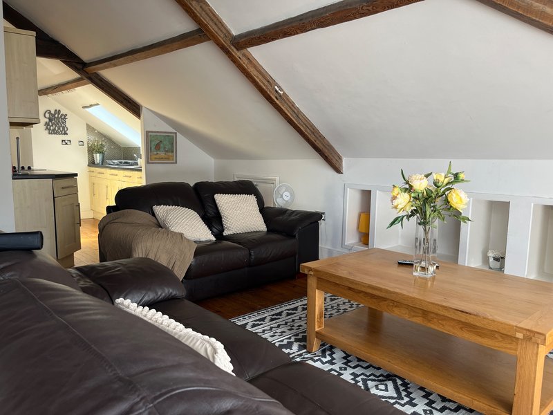 Open-plan attic living room with black sofas, wooden beams, yellow flowers in vase, and wooden coffee table