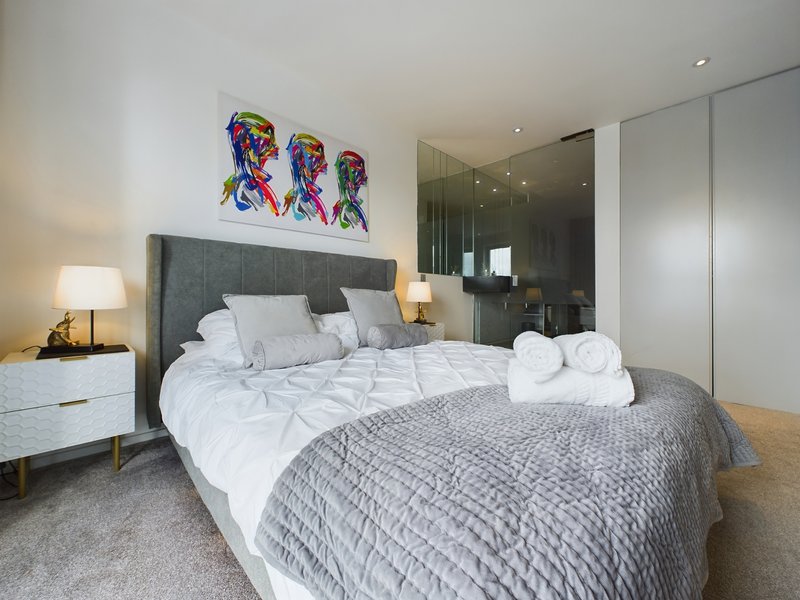 Modern bedroom with grey upholstered bed, white bedding, colorful abstract wall art, and ensuite bathroom visible