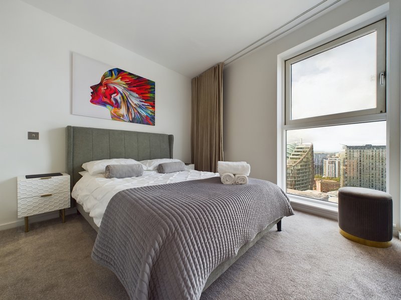 Modern bedroom with gray upholstered bed, large window with city views, and colorful wall art