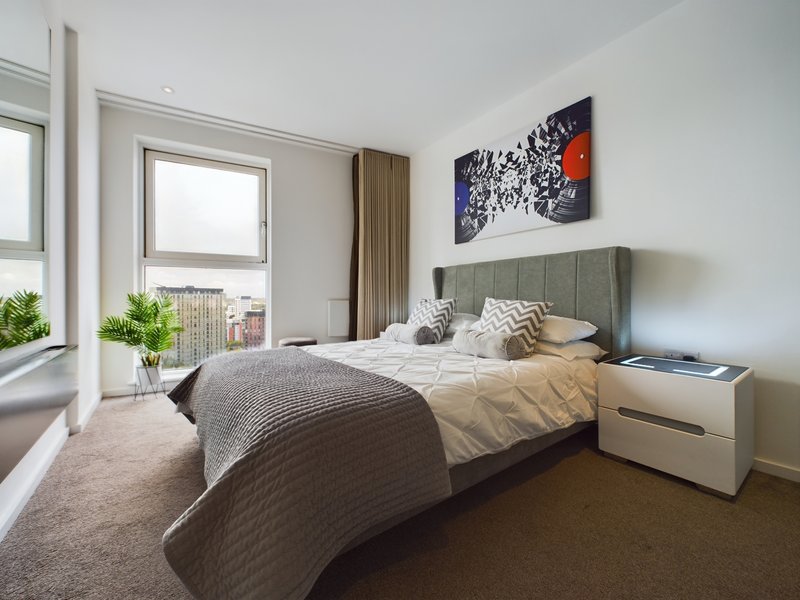 Modern bedroom with double bed, grey headboard, bedside table, and city view from windows