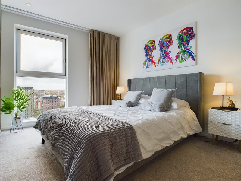 Modern bedroom with grey upholstered bed, white bedding, colorful wall art, and city view from window