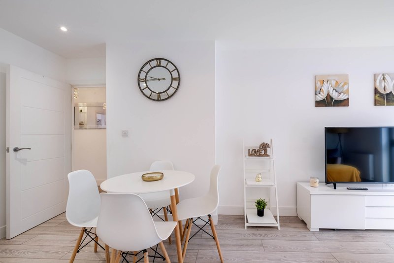 Modern living room with round white dining table, two chairs, wall clock, TV, and shelving