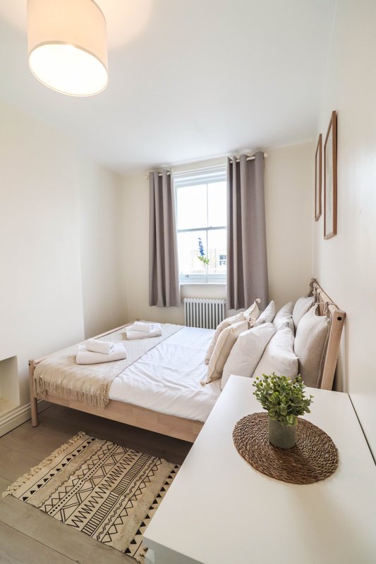 Bright bedroom with double bed, neutral bedding, window with curtains, potted plant on side table