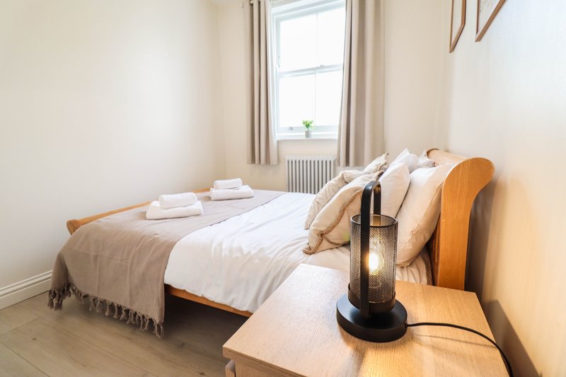 Bright bedroom with wooden bed frame, white bedding, cushions, and bedside lamp by window